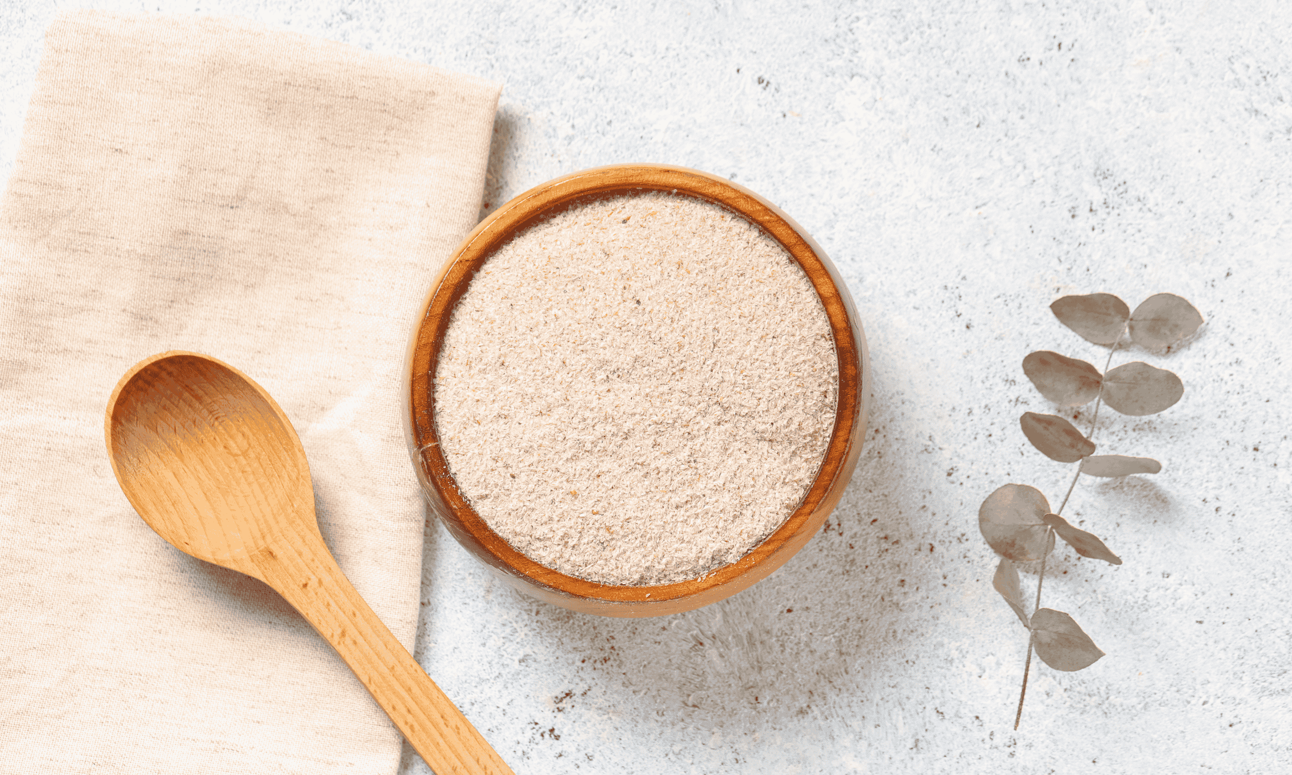 Draufsicht auf eine Holzschale gefüllt mit beigem Pulver oder Mehl, daneben ein Holzlöffel und Eukalyptuszweig.