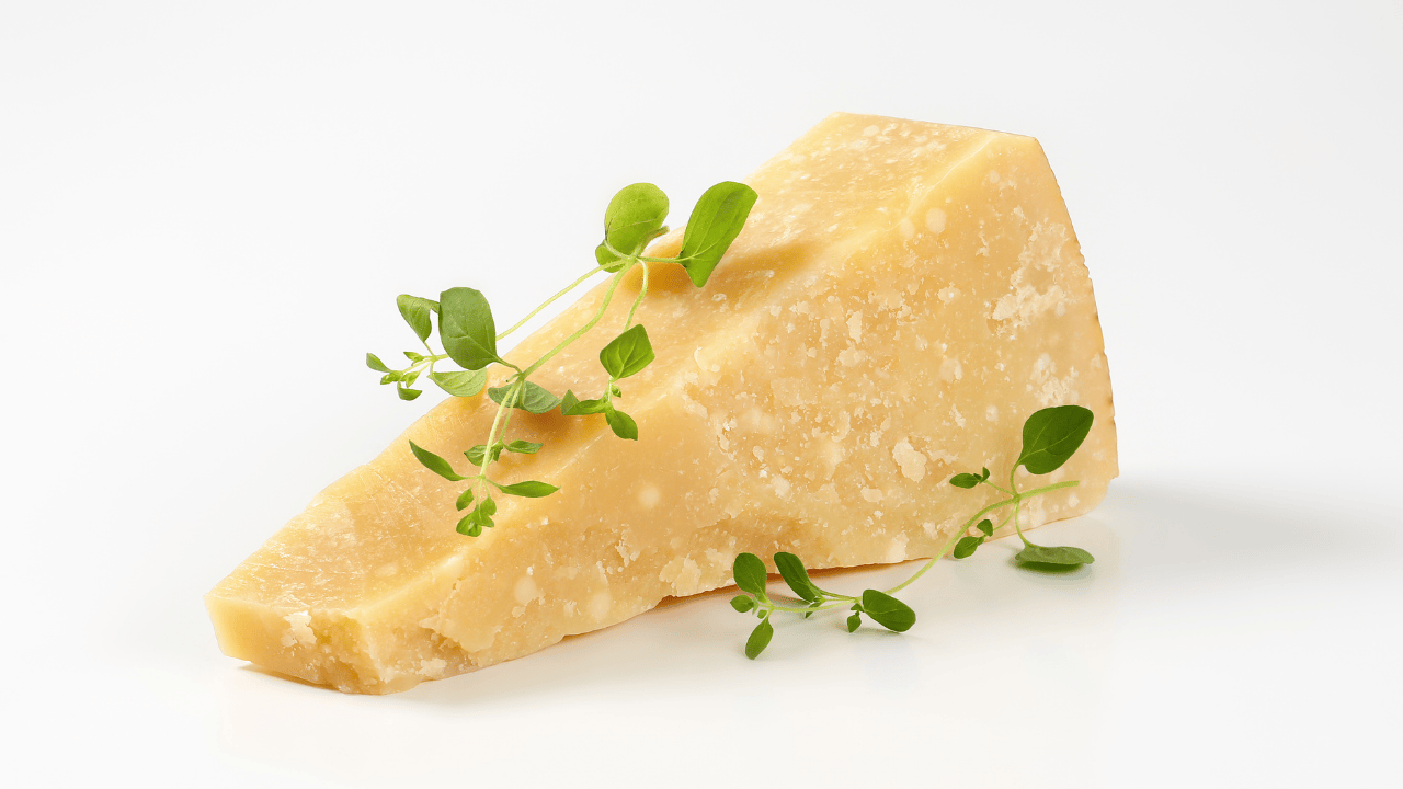 Stück Hartkäse auf weißem Hintergrund, dekoriert mit kleinen grünen Kräutern; minimalistische Food-Aufnahme.