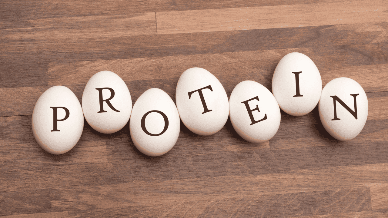 Weiße Eier auf Holzuntergrund, mit schwarzen Buchstaben beschriftet, ergeben das Wort „PROTEIN“.