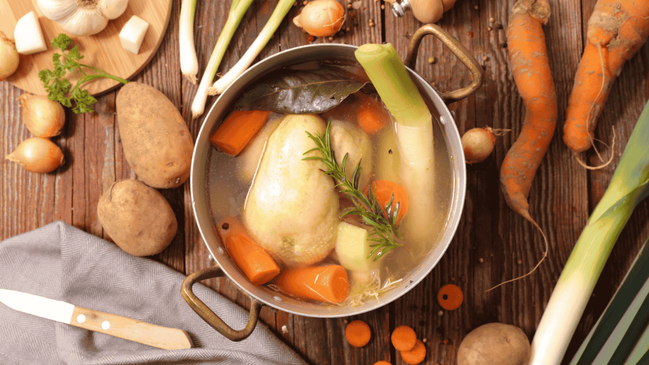 Topf mit Hühnerbrühe, Gemüse und Kräutern auf Holztisch, umgeben von frischen Zutaten.